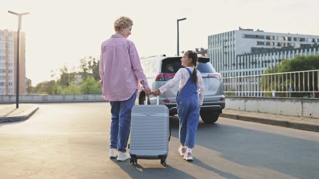 A Caucasian mother and her daughter are holding hands and pulling a suitcase next to a car, ready for a trip