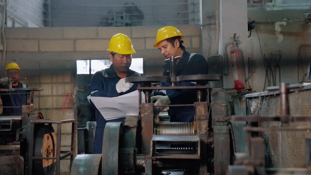 Industrial workers discussing machine operation and reviewing production plans beside heavy machinery in a manufacturing facility. Teamwork, communication, engineering, and workplace safety.
