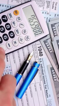 Calculating Income Tax Return. A hand enters figures on a calculator beside tax forms, dollar bills, and a blue pen.