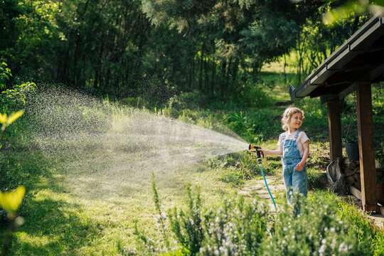 Happy little girl in denim overalls watering green lawn with garden hose on a sunny summer day