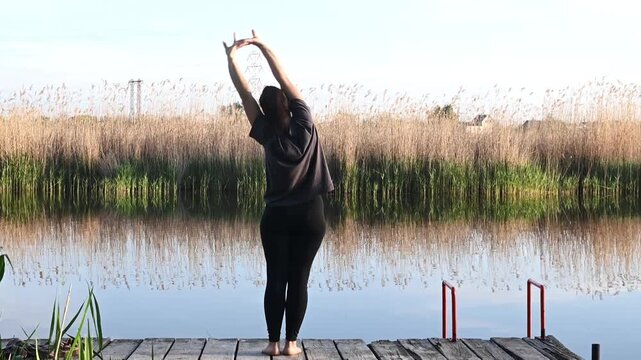Woman performs standing yoga stretch and bends with raised arms on wooden pier beside calm lake in natural setting. Practitioner improves flexibility and mindful awareness through wellness routine.
