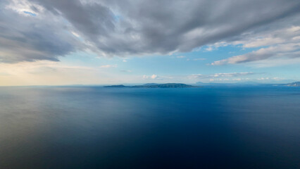 Storm clouds drifting above a tropical sea with a distant island on the horizon. Towering clouds contrast with calm blue water, creating a dramatic aerial seascape before changing tropical weather. © Houston