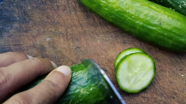 A close up of a person's hand using a knife to slice a fresh green cucumber on a wooden cutting board