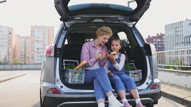 A Caucasian mother and her daughter smile while sitting in the open trunk of a car with groceries and donuts