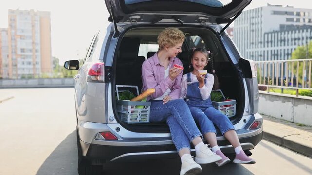 A Caucasian mother and her daughter enjoy donuts while sitting in the open trunk of a car after grocery shopping