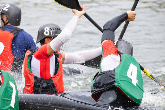 Canoe polo players in colorful jerseys engage in competitive action on a river, showcasing teamwork and skill with paddles raised and water splashing around them
