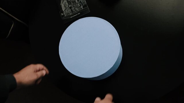 Two hands with dark sleeves set pale blue round hatbox onto black tabletop from close top view, then release it as box settles softly, creating gentle shadow, clean contrast, and calm commercial mood