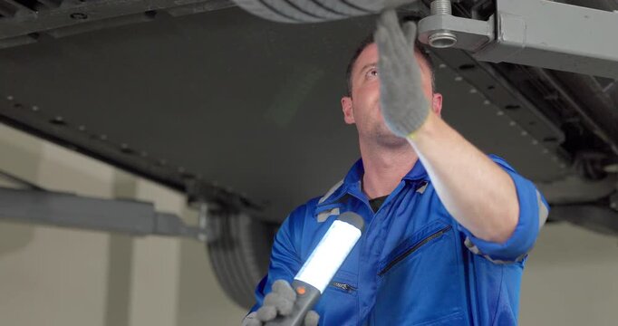 Mechanic inspecting car underbody with work light under vehicle on lift in modern garage