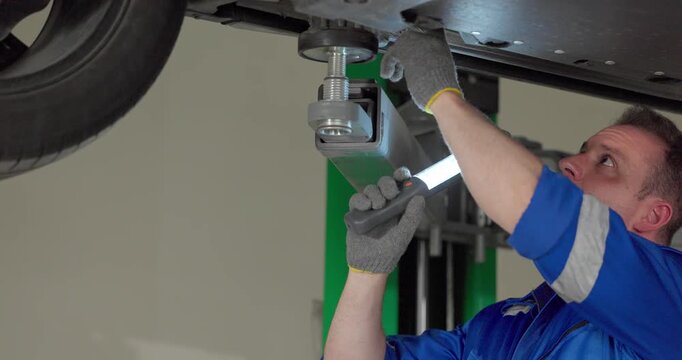 Mechanic inspecting car underbody with work light under vehicle on lift in modern garage