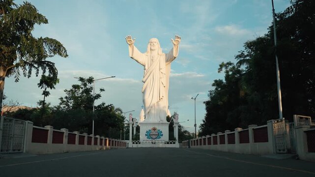 Cinematic gimbal push-in shot along a tree-lined road leading to the towering Sacred Heart of Jesus statue in Velankanni, arms raised against a soft morning sky in Tamil Nadu, India.