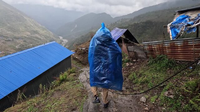 Hiker in blue poncho passes hillside lodges in the rain, Annapurna region, Nepal