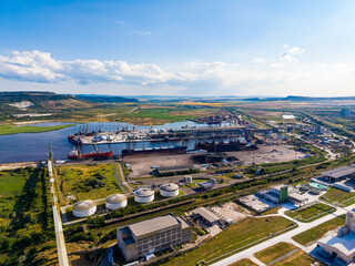Fototapeta premium View from above shows a large industrial site with cranes, ships, and storage tanks near a river and open land during the daytime