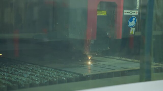 Laser cutting head producing bright molten spot on flat metal sheet inside enclosed industrial machine with slat bed, safety panel, glass reflection, precision fabrication details visible
