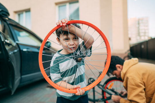 Boy holds orange bicycle wheel while father repairs bike outdoors