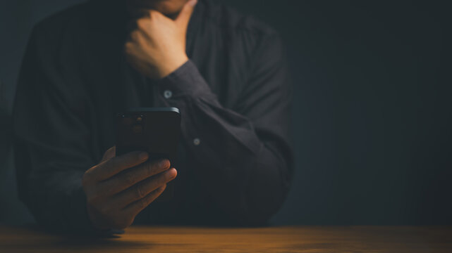 Serious business man thinking while holding a mobile phone in a dark room. Modern smartphone technology concept.