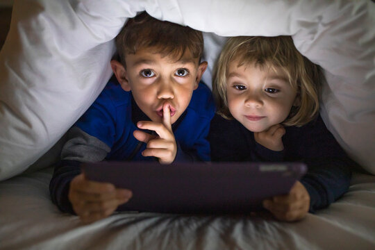 Portrait of brother and little sister lying side by side under a blanket using digital tablet