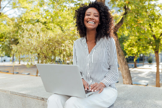 Curly haired businesswoman laughing while sitting with laptop at office park
