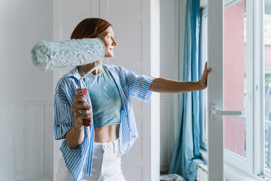 Smiling woman holds paint roller near window in new apartment