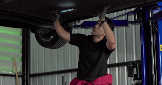 Mechanic inspecting car underbody with work light under vehicle on lift in modern garage