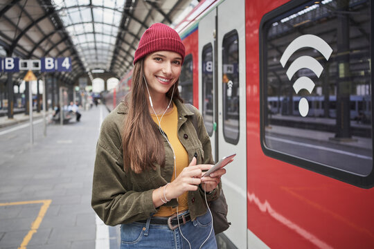 Smiling young female passenger with smart phone listening music through smart phone on railroad station