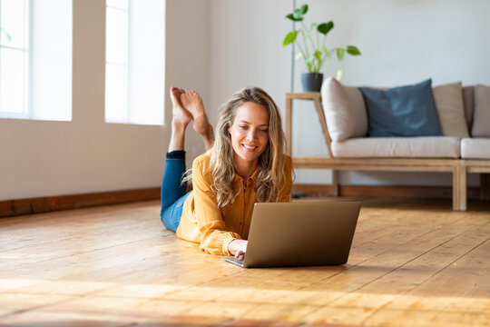 Smiling woman using laptop while lying on harwood floor in living room