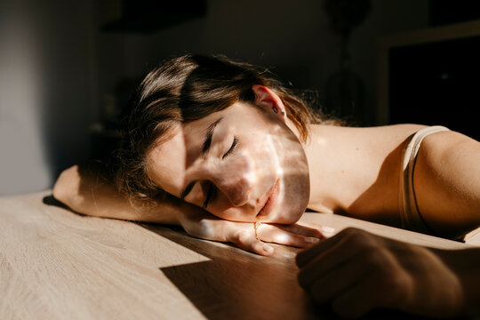 Portrait of young woman with eyes closed leaning on wooden table at sunlight