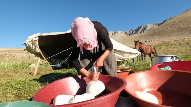 A woman squeezes a cloth bag of fresh curds over a red basin, showing a traditional cheese-making stage, manual whey draining, rural food preparation, and small-scale outdoor dairy production by hand.
