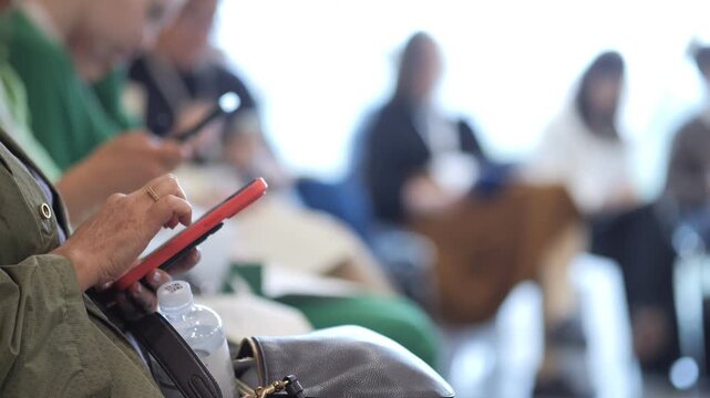 Person holds red tablet in public seating area. Background shows blurred attendees in conference or event space. Focus on device usage during gathering or meeting. Suitable for tech, business