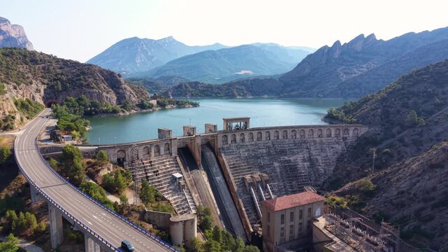 Aerial view of Oliana dam on the Segre River with elevated highway bridge curving through valley, turquoise reservoir and jagged mountain peaks, Lleida, Catalonia, Spain
