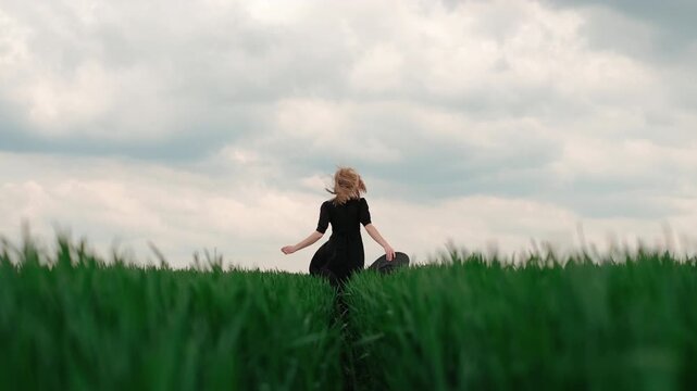Rear view shows young woman moving along narrow path through emerald grass, carrying wide brimmed hat in one hand while dark hair falls down back beneath soft overcast sky, with woman, walking, hat