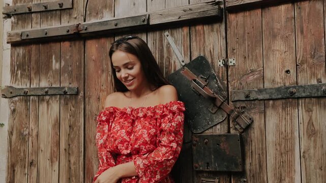 Young woman in red floral off-shoulder dress stands beside weathered wooden gate with iron hardware, smiling softly while making subtle pose changes, turning head, lowering hands, and crossing arms