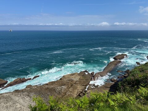 Panoramic view of a wild Atlantic coastline with ocean waves breaking against rugged rock formations beneath a blue sky. White sea foam and deep blue water capture the natural beauty of Cantabria, Spa