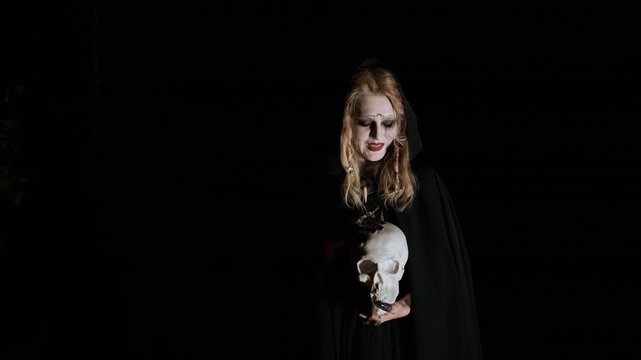Hooded pale woman with long blonde hair grips aged human skull at chest height, raising face upward and crying out under strong spotlight, creating dark gothic mood with eerie contrast