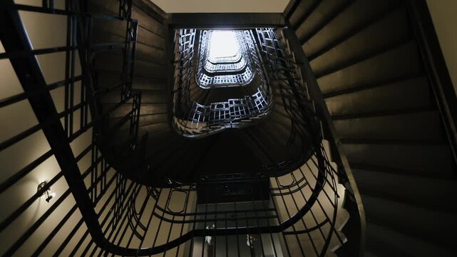Extreme low angle interior view looking straight up through rectangular spiral staircase, showing ornate wrought iron railings, stacked landings, bright skylight above, warm wall lighting, strong
