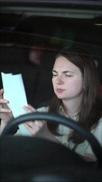Brunette woman seated in car interior studies a paper bill or receipt closely with focused worried expression while holding it in hand.