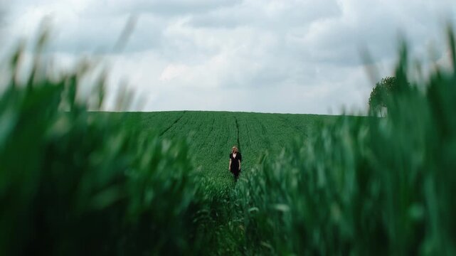 Lone person walking narrow path through tall emerald field under overcast sky, framed by soft blurred grass in foreground, creating tunnel-like leading lines, quiet rural mood, depth, and spacious