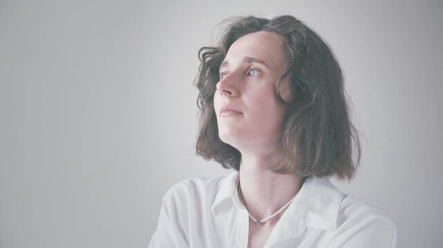 Close-up portrait of a young woman with curly black hair in thoughtful pose, resting chin on hand against soft neutral background with bright diffused lighting.