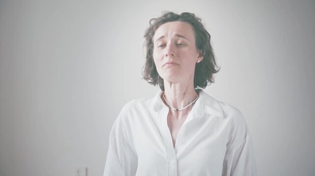 Curly haired young woman shaking head as hair flies, adjusting delicate necklace indoors in white collared shirt, close up portrait with hands and thoughtful expression