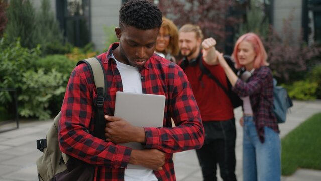 Upset African American guy male student bullied man feeling despair bullying by multiracial students bad gossiping showing thumb down judge disrespect teasing mocking bully at city college university
