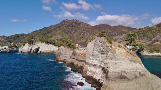 Dramatic drone shot flying over unique stratified sedimentary rock formations along the beautiful blue coastline of the Izu Peninsula, Japan.