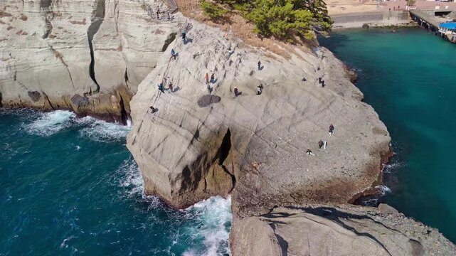 High angle drone footage showing the unique layered tuff rock structures and tourists walking by the turquoise sea in Dogashima, Izu.