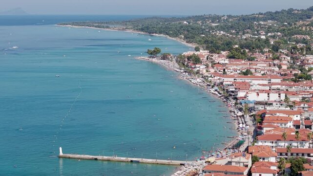 Crowded Pefkochori beach and coastal resort town on a sunny day, beachfront hotels, cafe, villas with mediterranean greenery, Halkidiki, Kassandra peninsula, Aegean Sea, Aerial view