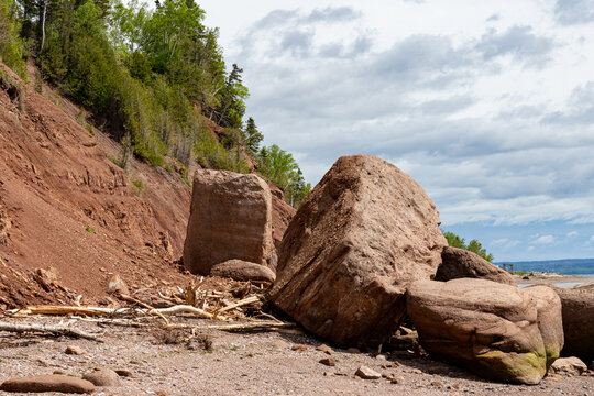 Gaspesie, Quebec, 07 June 2026 : Large red boulders and driftwood lie on a rocky beach beneath a steep, eroded clay cliff.