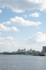 Obraz premium Vertical landscape of a wide river featuring a cargo barge, soaring birds, and a residential cityscape under a bright blue sky. Great for urban lifestyle imagery and riverfront backgrounds.