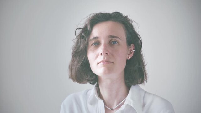 Young woman sitting upright in white shirt against gray studio background, showcasing neutral to warm expressions for lifestyle and wellness themes.