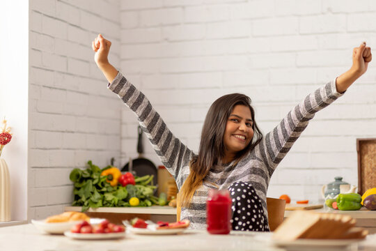Cheerful woman sitting with arms raised in kitchen during breakfast