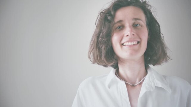 Smiling woman with short curly hair in white shirt against soft gray background, showcasing beauty and wellness in soft diffused lighting.
