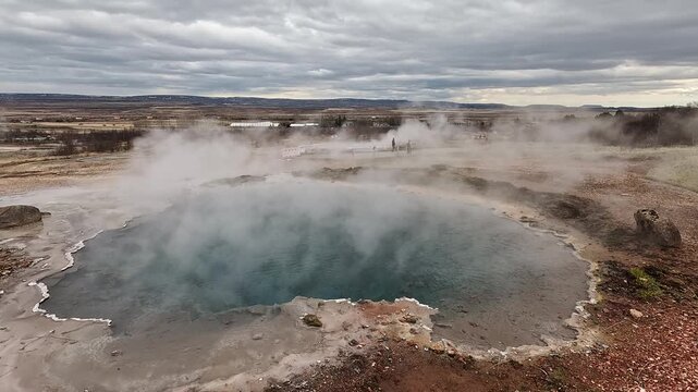 Strokkur geyser steaming silica terrace pool wide shot with Haukadalur Valley farmland beyond, Iceland