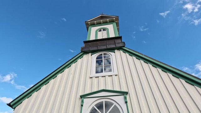 Low-angle upward shot of Thingvallakirkja historic church tower with cream and green facade against blue sky, Iceland