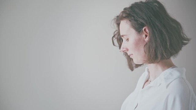 Young woman in profile with shoulder-length hair and white shirt, eyes closed, in calm indoor setting, soft neutral background, conveying mindfulness and introspection.